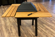 A perspective view of the entire table. The black grill insert is in the center, now covered with a flat, black lid, and surrounded by an ample wooden slatted surface. The sturdy black legs support the table, which can serve as both a grill and a dining area.