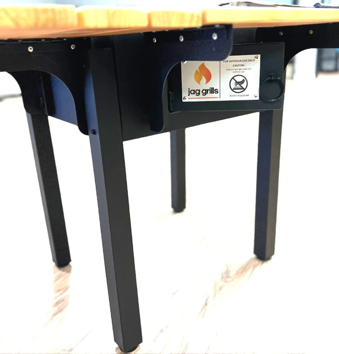 A side view of the table showcasing its black metal frame and legs. A label with the logo 'jag grills' is visible on one side, indicating the brand and containing warning information such as "FOR OUTDOOR USE ONLY" and safety certifications.