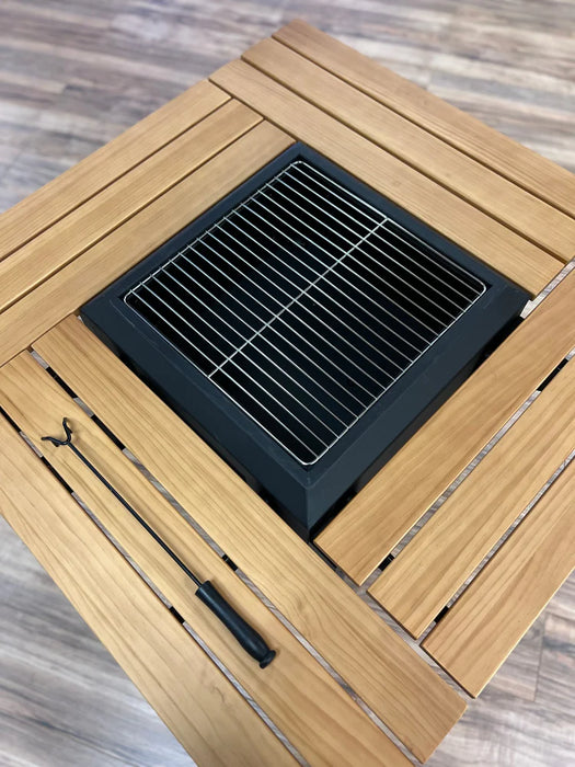 A close-up overhead view of the same table, now showing the square grill insert covered with a stainless steel cooking grate. The grate has a grid pattern suitable for grilling. The long black poker and a small black handle sit neatly on the wooden slats.