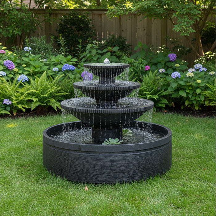 Three tier black garden fountain with wide round basin placed on green lawn bordered by hydrangeas and backyard fence