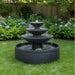Three tier black garden fountain with wide round basin placed on green lawn bordered by hydrangeas and backyard fence