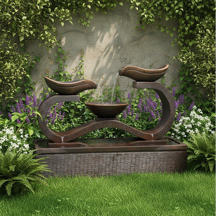 Tiered water fountain, Exaco Infinity in Rust finish, surrounded by ivy, purple flowers, and green lawn.