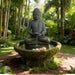Tropical stone Buddha bowl water fountain with cascading water, moss accents, and palm trees in serene garden setting.