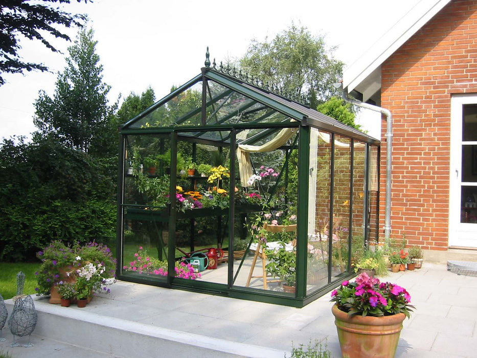 Victorian 8x10 greenhouse positioned on a patio, filled with colorful potted plants and curtain shades, adjacent to a brick home.