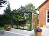 Victorian 8x10 greenhouse on patio with blooming flowers inside and out, showing the Exaco Janssens Royal Victorian VI 23’s elegant glass and frame.