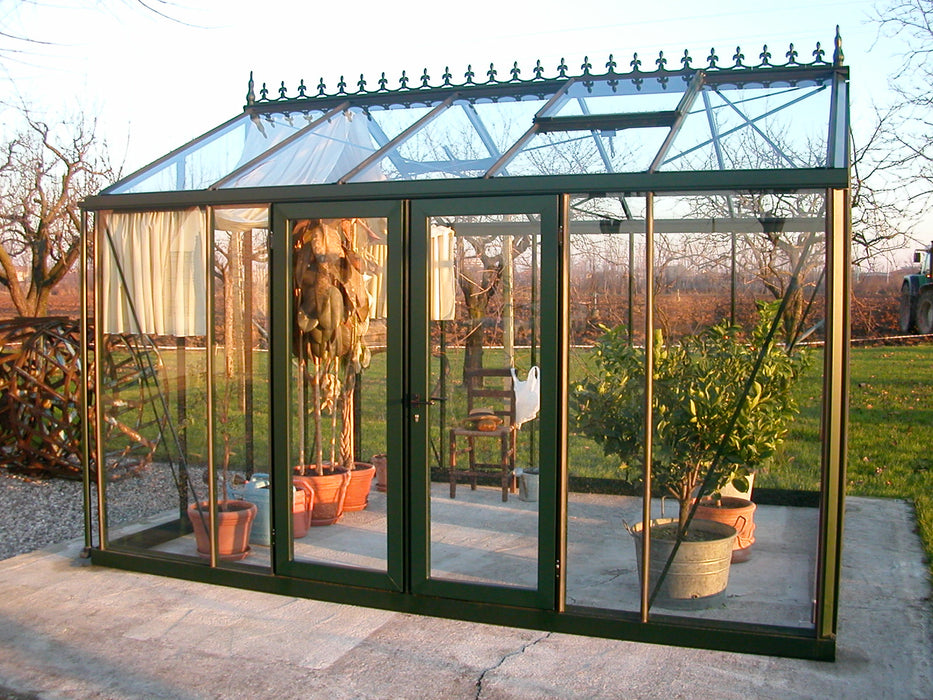 Victorian VI 23 greenhouse with double turning doors, sunlight streaming through its glass panels, and potted citrus plants in an open countryside backdrop.