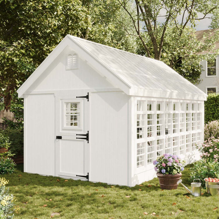 Little Cottage Co. Colonial Gable Greenhouse in a lush garden setting with white Dutch door, grid windows, and surrounding flowers.