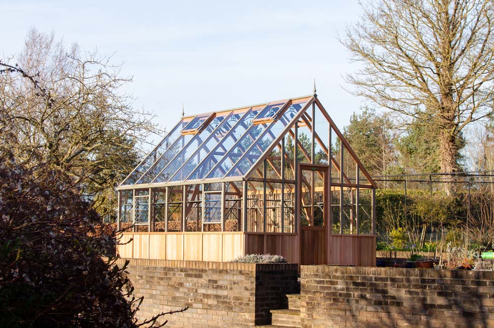 Wide shot of a cedar Westminster greenhouse positioned on a raised brick platform, surrounded by trees, greenery, and a bright outdoor setting with clear skies.