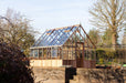 Wide shot of a cedar Westminster greenhouse positioned on a raised brick platform, surrounded by trees, greenery, and a bright outdoor setting with clear skies.