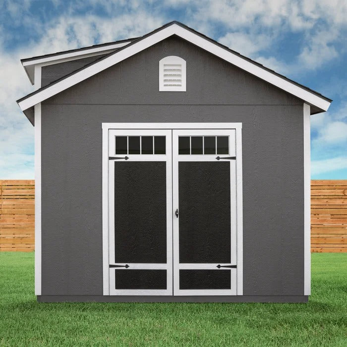 A Handy Home Windemere 10x12 - Wood Storage Shed, in dark gray with white trim, features double doors and a small attic window. Set against a wooden fence under a partly cloudy sky, this weather-resistant structure offers ample storage for all your needs.
