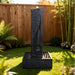 Modern outdoor water fountain in black stone finish with textured column and square basin, displayed in a landscaped backyard garden.