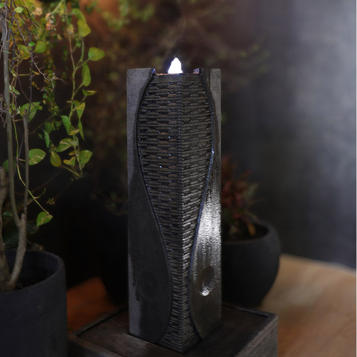 Yang modern tabletop water fountain with vertical textured panel and soft LED glow, styled indoors with plants for cozy ambiance.