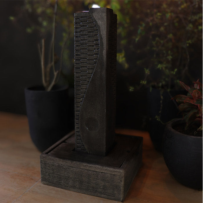 Side view of Yang sculptural outdoor water fountain with curved stone design and square base, styled among potted plants at night.