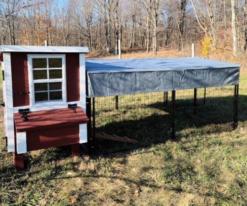 A small red OverEZ chicken coop with white trim and windows, adjacent to a covered outdoor chicken run in a grassy area, part of the OverEZ Small Flock Bundle Basic.