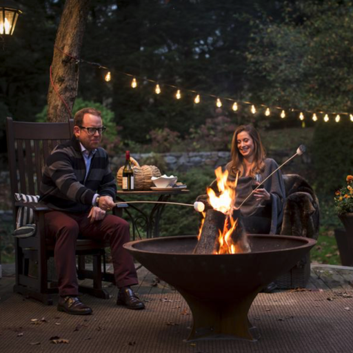 A couple enjoying a cozy evening by a lit Arteflame 40 inch fire pit with a low euro base, roasting marshmallows and savoring a relaxed outdoor moment.