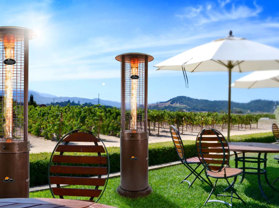 A tall, bronze outdoor flame heater Vulcan stands on a garden beside a dining area adding comfort and elegance to the garden scene.
