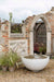 The Solus Decor Water Dome in Halva white is nestled in a classic garden setting, framed by an elegant stone archway, blending the tranquility of water with the grandeur of historical architecture.