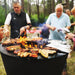 Outdoor cookout with a group of people grilling various meats on a 40-inch Arteflame One Series grill in a forest setting.
