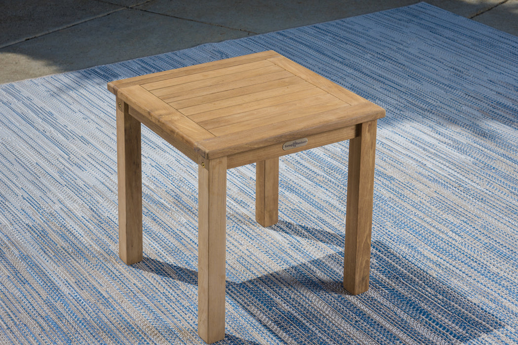 A small Tortuga Outdoor teak side table on a blue rug, featuring all-weather resin wicker construction.