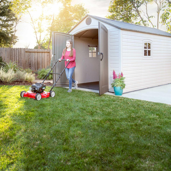 A woman is mowing the lawn in front of a Lifetime 8 Ft. X 15 Ft. Outdoor Storage Shed - 60075.