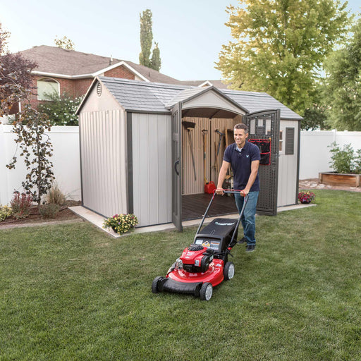 A man is mowing his lawn in front of a Lifetime 12.5 Ft. X 8 Ft. Outdoor Storage Shed - 60223.
