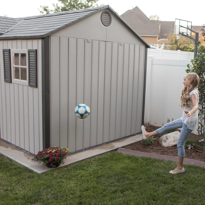 A girl kicking a soccer ball in front of a Lifetime 12.5 Ft. X 8 Ft. Outdoor Storage Shed - 60223 shed.