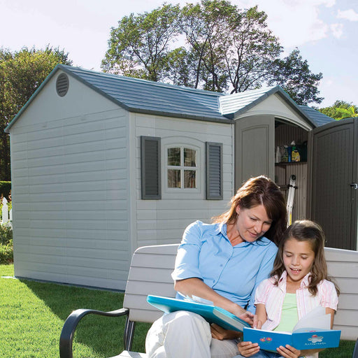 A woman and a girl sitting outside a Lifetime 15 Ft. X 8 Ft. Outdoor Storage Shed - 6446.