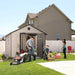 A family is mowing the lawn in front of a Lifetime 11 Ft. X 13.5 Ft. Outdoor Storage Shed - 6415.