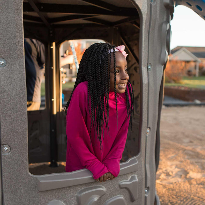 A smiling girl with braided hair peeks out from the playhouse section of the grey and blue Lifetime Adventure Tower playset.