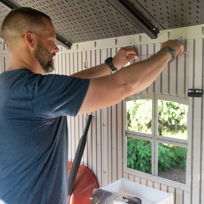 A man is putting screws into the wall of a Lifetime 10 Ft. X 8 Ft. Outdoor Storage Shed - 60005.