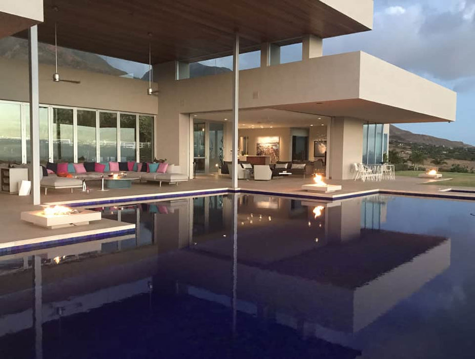 A modern home features the Solus Decor Halo Low Fire Pit near an infinity pool, reflecting flames in the water for a dramatic evening setting.