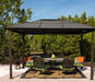 Paragon Outdoor Madrid Hard Top Gazebo on a patio with a full dining set and green and orange accents, set against a backdrop of lush greenery.