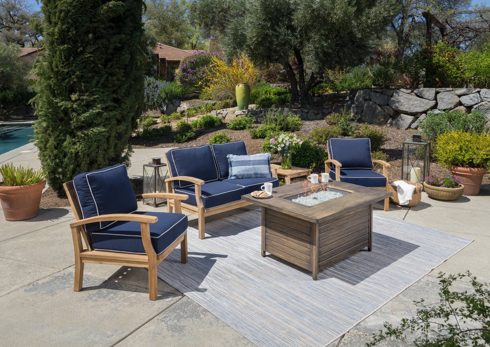 This luxurious Tortuga Outdoor 5-Piece Indonesian Teak Loveseat and Fire Table Set - Canvas Natural or Navy features outdoor wicker furniture adorned with vibrant blue cushions and a matching rug, creating the perfect retreat for your outdoor space.